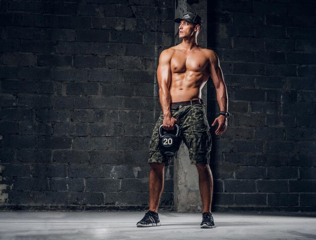 El hombre sin camisa enfocado con gorra está haciendo ejercicios con pesas en un estudio fotográfico oscuro.