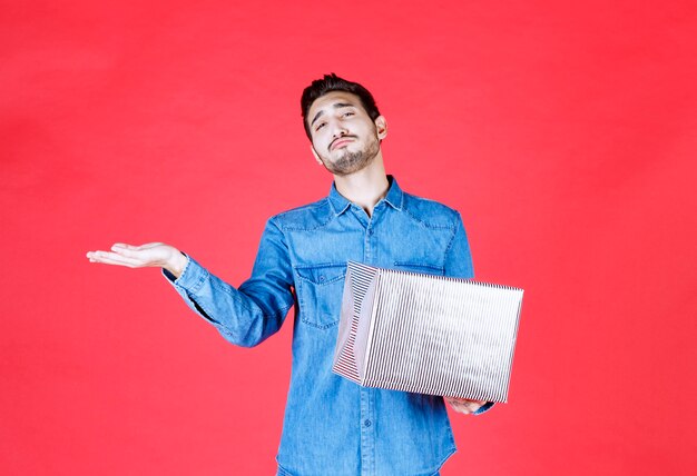 Hombre en camisa de mezclilla con caja de regalo plateada y apuntando a algún lugar.
