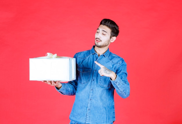 Hombre en camisa de mezclilla con caja de regalo plateada y apuntando a algún lugar.