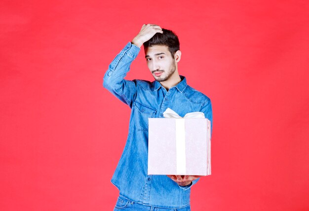 Hombre en camisa de mezclilla sosteniendo una caja de regalo púrpura atada con cinta blanca y parece confundido y pensativo.