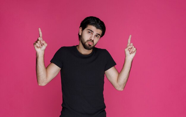 Hombre de camisa negra con barba apuntando a algo de manera confusa o mostrando la dirección