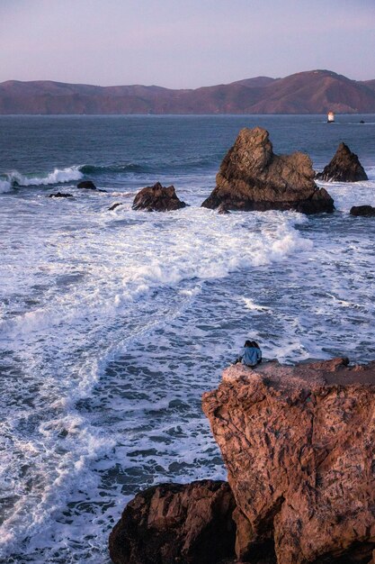 Hombre con camisa negra sentado en la formación de roca marrón cerca del mar durante el día