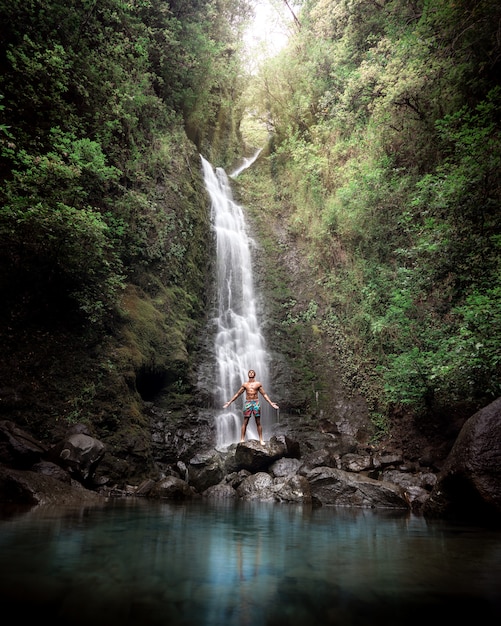 Hombre sin camisa de pie sobre las rocas cerca de una hermosa cascada con un lago y vegetación