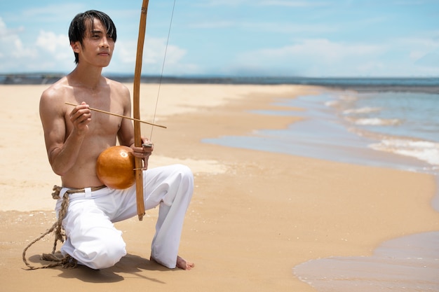 Foto gratuita hombre sin camisa practicando capoeira en la playa con arco de madera