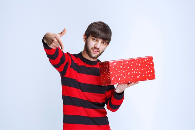 Hombre con camisa a rayas rojas con una caja de regalo roja y llamando a alguien para que se la presente.