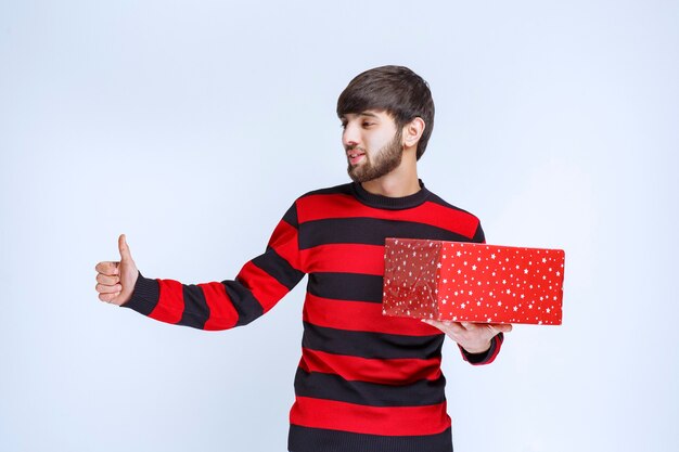 Hombre en camisa de rayas rojas con una caja de regalo roja y mostrando signo de disfrute.