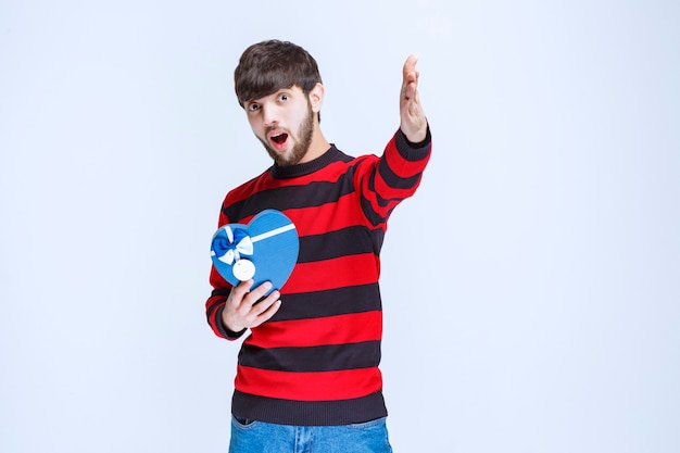 Hombre con camisa de rayas rojas sosteniendo una caja de regalo con forma de corazón azul y llamando o notando a alguien más adelante.