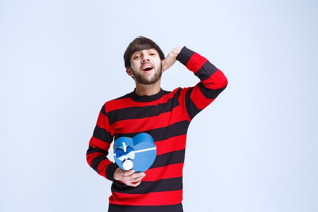 Hombre con camisa de rayas rojas sosteniendo una caja de regalo con forma de corazón azul y llamando o notando a alguien más adelante.