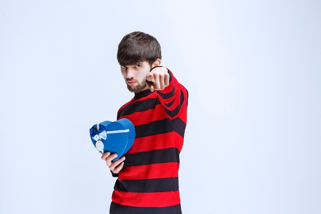 Hombre con camisa de rayas rojas sosteniendo una caja de regalo con forma de corazón azul y llamando o notando a alguien más adelante.