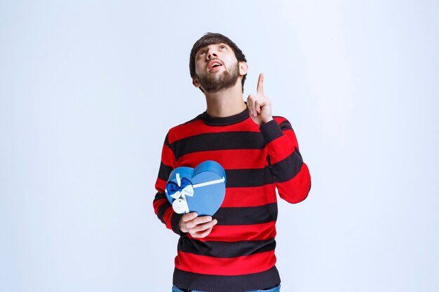 Hombre con camisa de rayas rojas sosteniendo una caja de regalo con forma de corazón azul y parece pensativo o como si tuviera una buena idea.