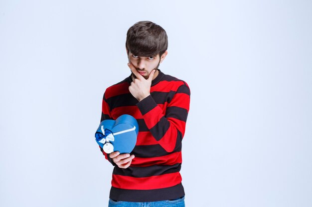 Hombre con camisa de rayas rojas sosteniendo una caja de regalo con forma de corazón azul y parece pensativo o como si tuviera una buena idea.