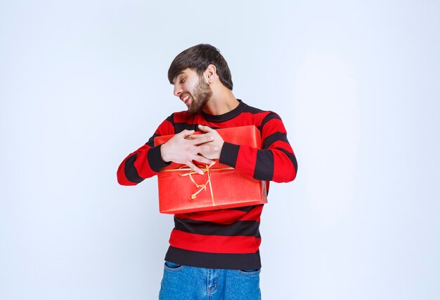 Hombre con camisa de rayas rojas sosteniendo una caja de regalo roja y abrazándola fuerte y no quiere compartir con nadie.