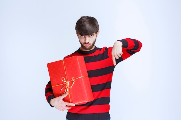 Hombre con camisa a rayas rojas sosteniendo una caja de regalo roja y llamando a la persona que está junto a él.