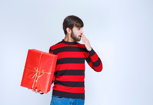 El hombre con camisa de rayas rojas sosteniendo una caja de regalo roja parece cansado y con sueño.