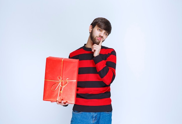 El hombre con camisa de rayas rojas sosteniendo una caja de regalo roja y parece confundido y pensativo.