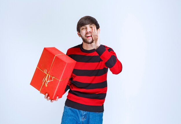 Hombre con camisa de rayas rojas sosteniendo una caja de regalo roja y pidiendo que alguien la entregue.