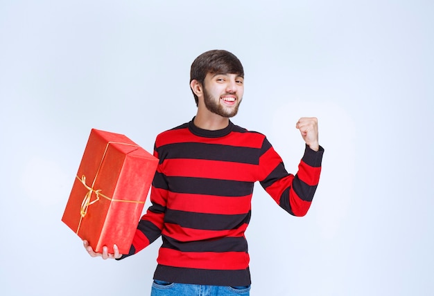 Hombre con camisa de rayas rojas sosteniendo una caja de regalo roja y sintiéndose poderoso y positivo.