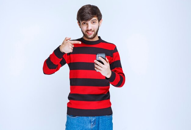 Hombre con camisa de rayas rojas sosteniendo un teléfono inteligente negro y apuntando a él.