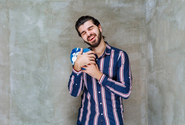 Hombre con camisa a rayas sosteniendo una caja de regalo con forma de corazón azul y abrazándola como se siente muy feliz