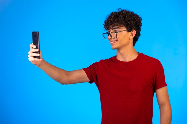 Hombre de camisa roja tomando selfie o haciendo una llamada telefónica y divirtiéndose sobre fondo azul.