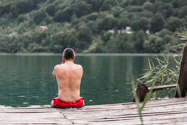 Hombre sin camisa sentado en el muelle de madera cerca del lago