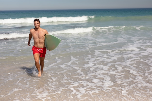 Hombre sin camisa con tabla de surf caminando en la playa bajo el sol