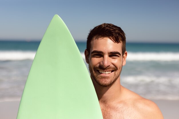 Hombre sin camisa con tabla de surf mirando a la cámara en la playa bajo el sol