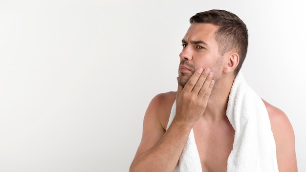 Hombre sin camisa con una toalla alrededor de su cuello comprobando la cara de pie contra el fondo blanco.