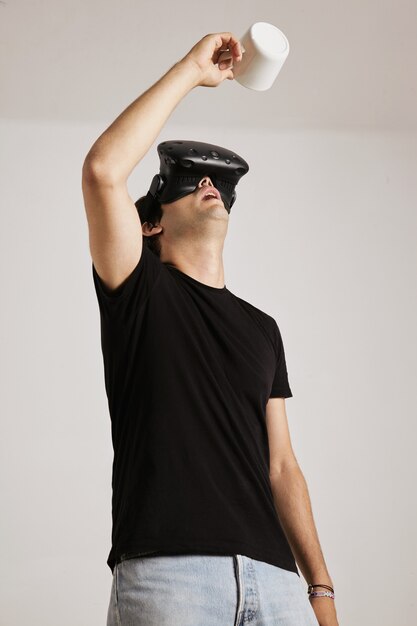 Un hombre con camiseta negra en blanco y casco de realidad virtual está mirando hacia una taza blanca vacía que sostiene sobre su cabeza, aislado en blanco