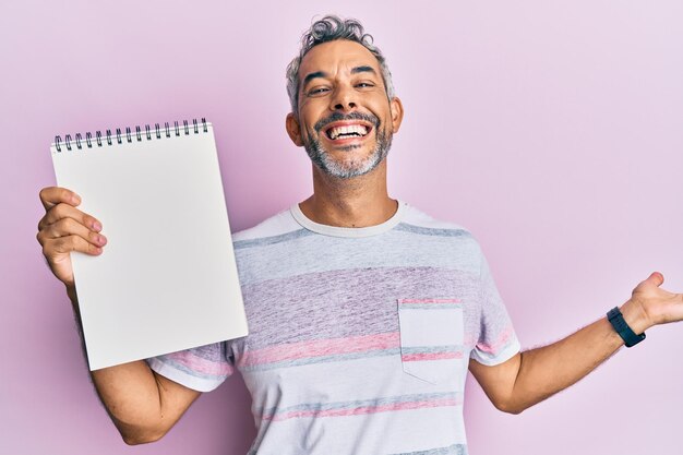 Hombre canoso de mediana edad sosteniendo un cuaderno vacío celebrando la victoria con una sonrisa feliz y expresión ganadora con las manos levantadas