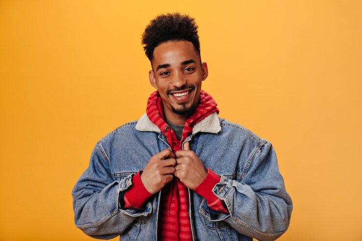 Foto gratuito hombre con capucha abotonándose la chaqueta de mezclilla y posando sobre fondo naranja chico encantador con sudadera roja sonriendo ampliamente sobre un fondo aislado