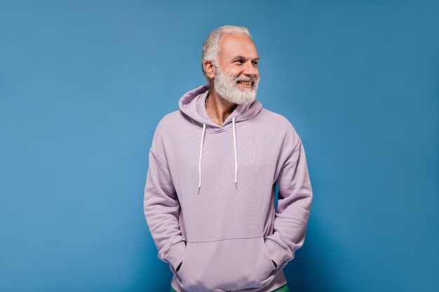 Hombre con capucha gris posando sobre fondo azul Chico adulto alegre con barba blanca en sudadera lila mirando hacia otro lado y sonriendo