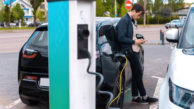 Hombre cargando su coche eléctrico en la estación de carga y usando smartphone