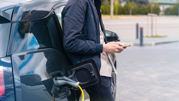 Hombre cargando su coche eléctrico en la estación de carga y usando smartphone