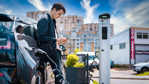 Hombre cargando su coche eléctrico en la estación de carga y usando smartphone