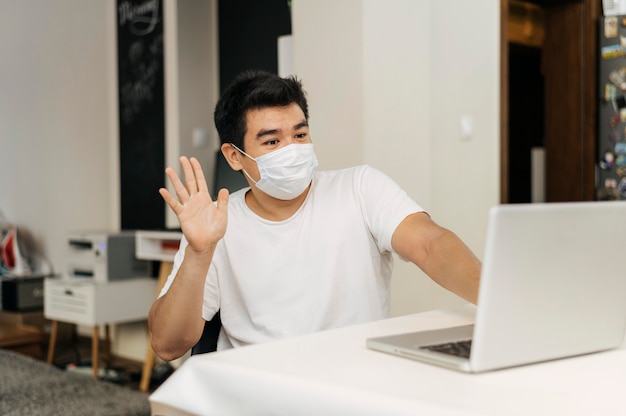 Hombre en casa con máscara médica durante la pandemia saludando a la computadora portátil