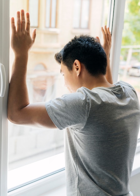 Hombre en casa durante la pandemia apoyando los brazos contra la ventana