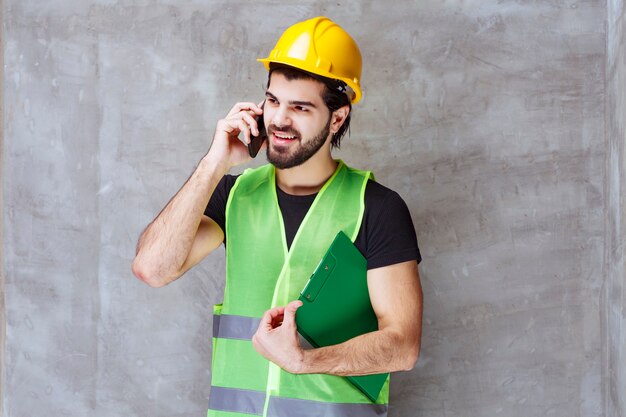 Hombre con casco amarillo y equipo sosteniendo una carpeta de informes y hablando por teléfono