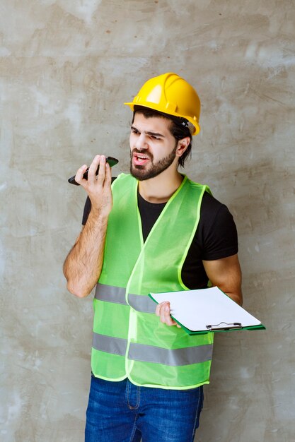 Hombre con casco amarillo y equipo sosteniendo un plan de proyecto y hablando por teléfono