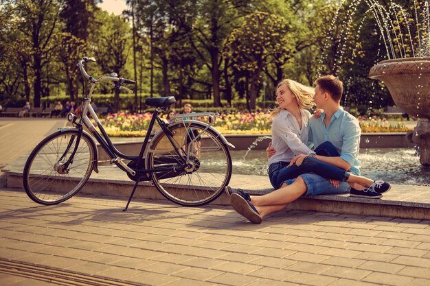 Hombre casual con bicicleta besando a su novia cerca de una fuente en un parque.