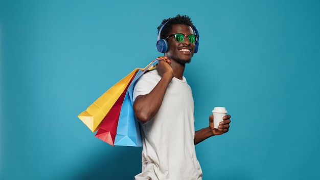 Hombre casual que lleva bolsas de compras y usa auriculares. Persona moderna escuchando música mientras sostiene bolsos con ropa de la tienda minorista y una taza de café. adulto adicto a las compras