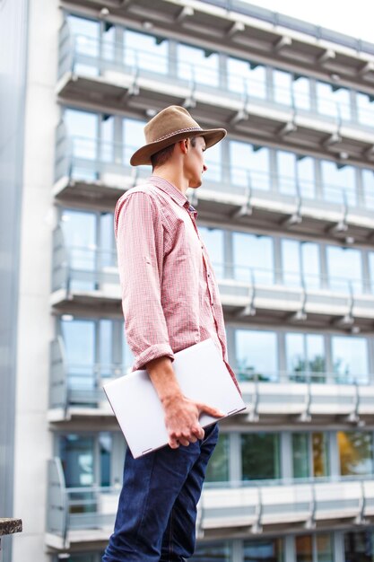 Hombre casual en un sombrero sosteniendo una computadora portátil en el techo contra un edificio de la ciudad.