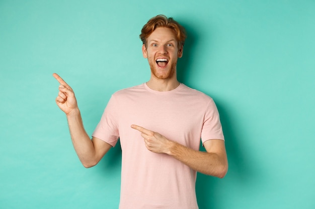 Hombre caucásico alegre en camiseta que parece feliz, señalando con el dedo hacia la izquierda y mostrando una oferta promocional, de pie sobre un fondo turquesa