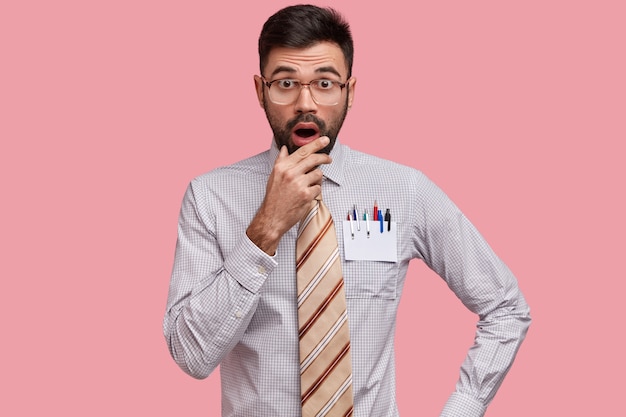 Hombre caucásico aterrorizado con barba oscura, mantiene la mano en el mentón, tiene expresión estupefacta, viste camisa formal y corbata a rayas