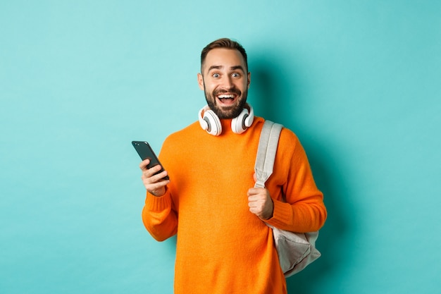 Hombre caucásico con auriculares y mochila sorprendido después de leer la notificación del teléfono