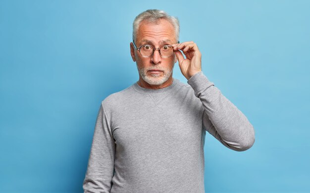 Hombre caucásico barbudo senior sorprendido que mira a través de anteojos expresa conmoción maravillas descuentos estacionales y precios escucha noticias increíbles usa jersey gris casual aislado sobre pared azul