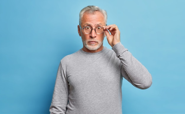 Hombre caucásico barbudo senior sorprendido que mira a través de anteojos expresa conmoción maravillas descuentos estacionales y precios escucha noticias increíbles usa jersey gris casual aislado sobre pared azul