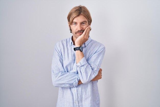 Hombre caucásico con bigote parado sobre fondo blanco pensando que parece cansado y aburrido con problemas de depresión con los brazos cruzados.