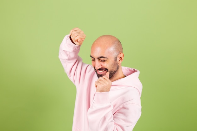 Hombre caucásico calvo joven en sudadera con capucha rosa aislado, feliz y alegre, sonriente movimiento casual y confiado escuchando música