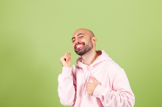 Hombre caucásico calvo joven en sudadera con capucha rosa aislado, feliz y alegre, sonriente movimiento casual y confiado escuchando música